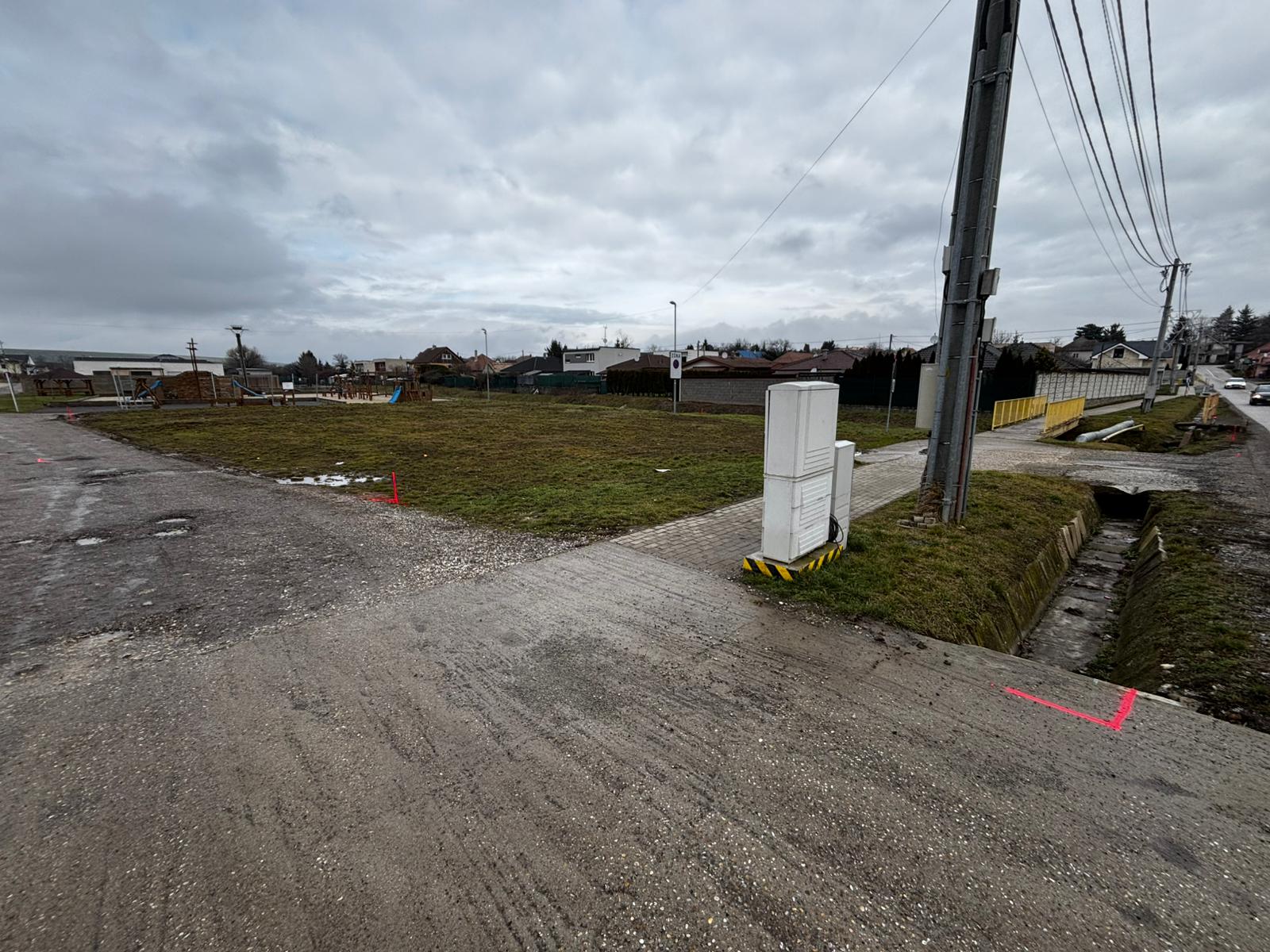 PRED — pôvodný stav pozemku v centre obce (~1 200 m²). Červené geodetické značky vyznačujú hranice budúcej stavby.