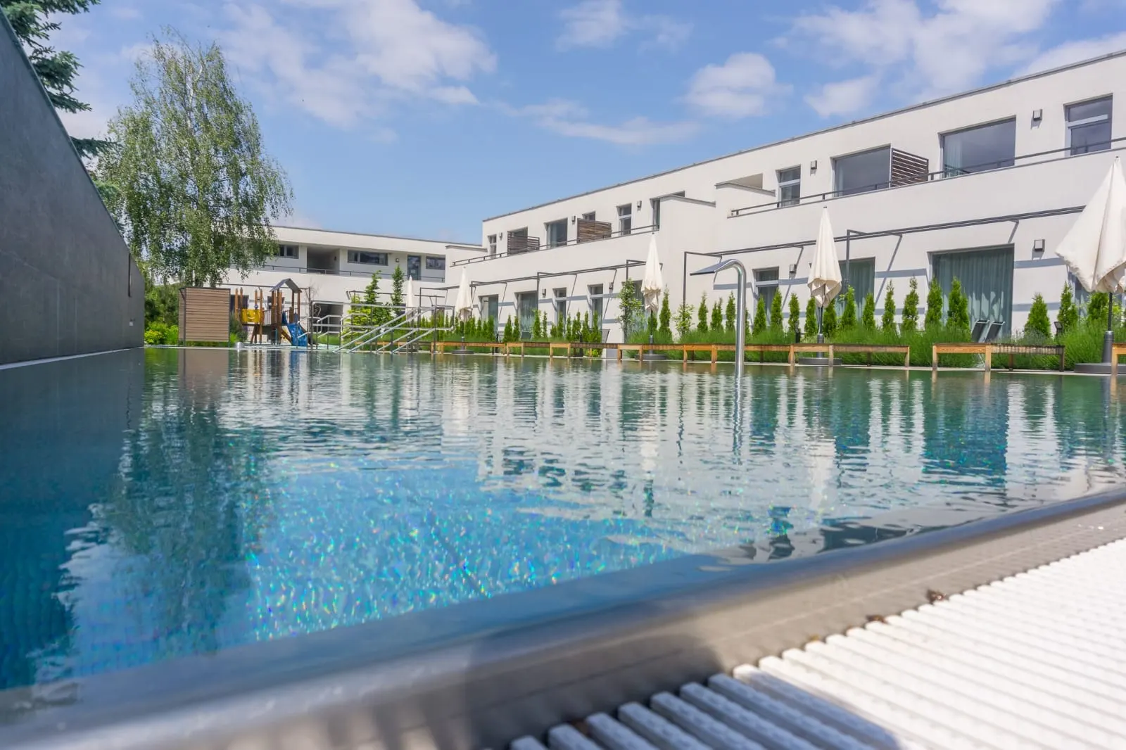 Vonkajší bazén v centre areálu — pohľad na ubytovaciu časť s balkónmi a zelenou zónou pred apartmánmi.