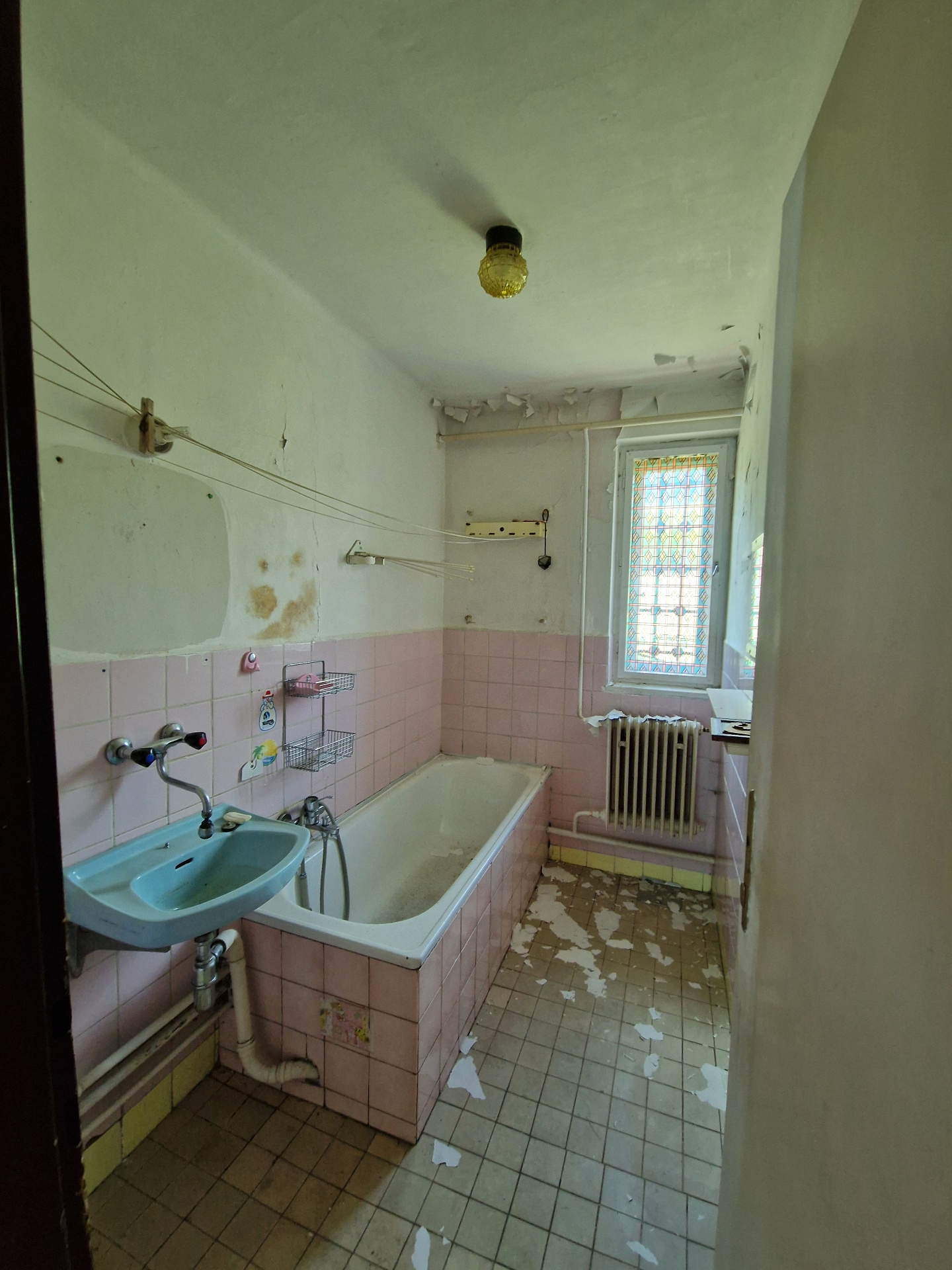 PRED rekonštrukciou (05/2025) — pôvodná kúpeľňa s ružovým obkladom, liatinovou vaňou, modrým umývadlom a vitrážovým oknom. Celé sociálne zázemie bude nahradené novým wellness-orientovaným riešením.