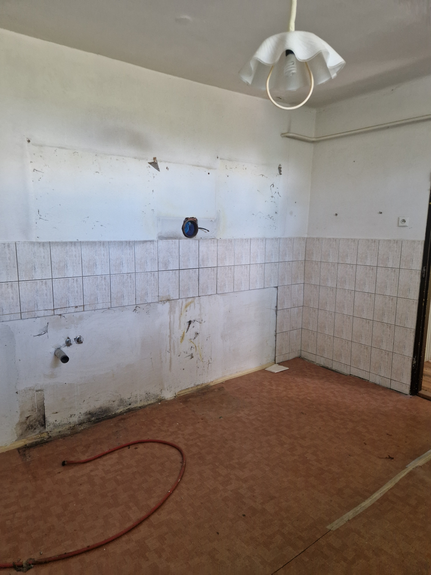 PRED rekonštrukciou (05/2025) — pôvodná kuchyňa po vypratení nábytku, s viditeľnými prípojkami vody, starým obkladom a PVC podlahou. Priestor je pripravený na demontáž a kompletnú obnovu.